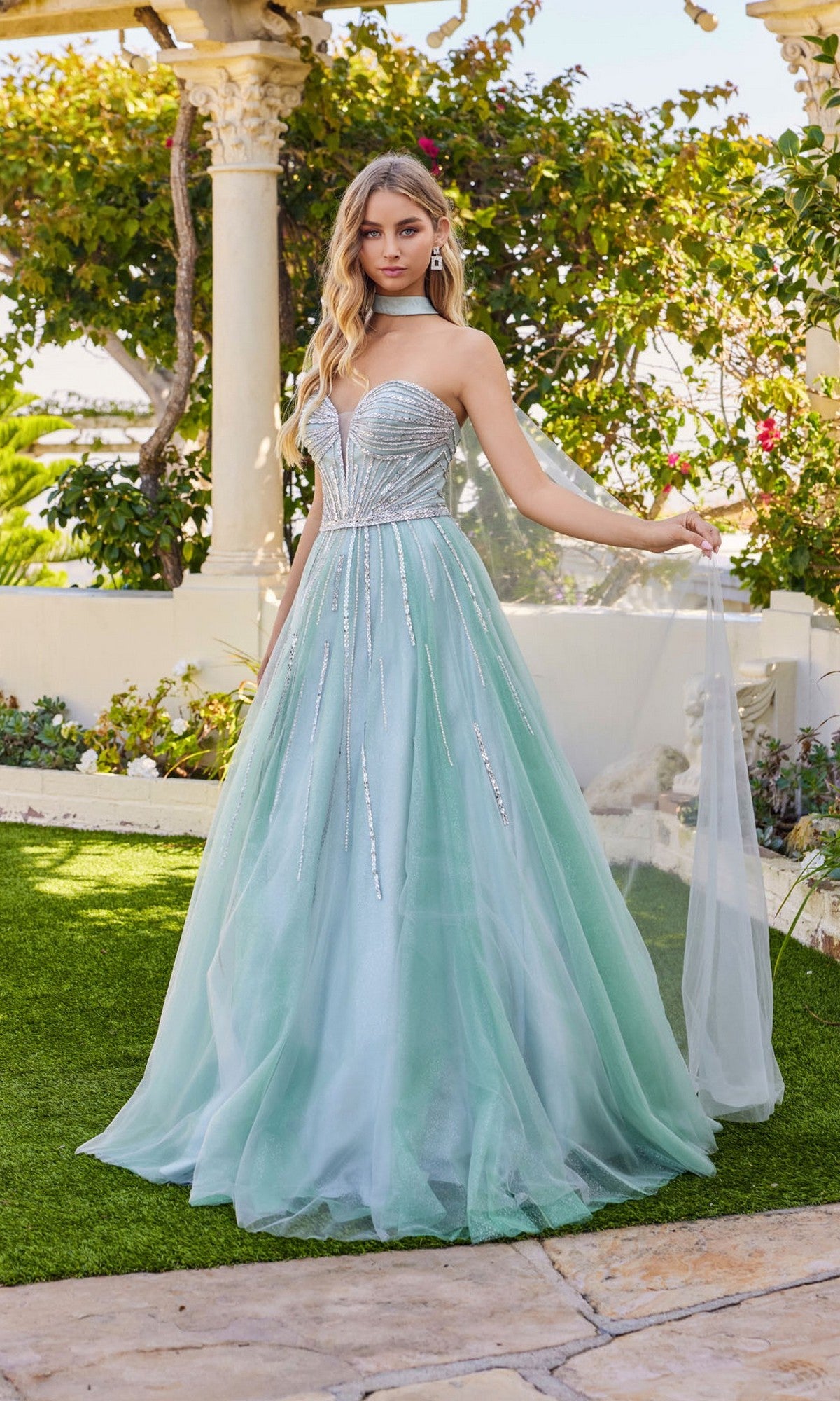 A woman stands outdoors wearing the Juliet JT2620U long prom dress in sage green, featuring a strapless, rhinestone-embellished bodice and a flowing tulle skirt.